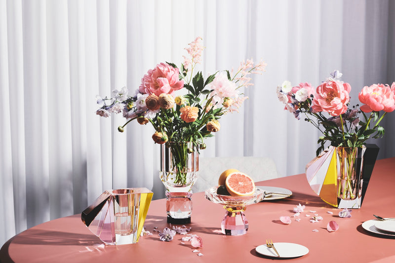Opsætning af vaser i klar, brun, gul og lyserød krystal med blomster i samt opsats/skål med frugter, på bord med koralfarvet dug.