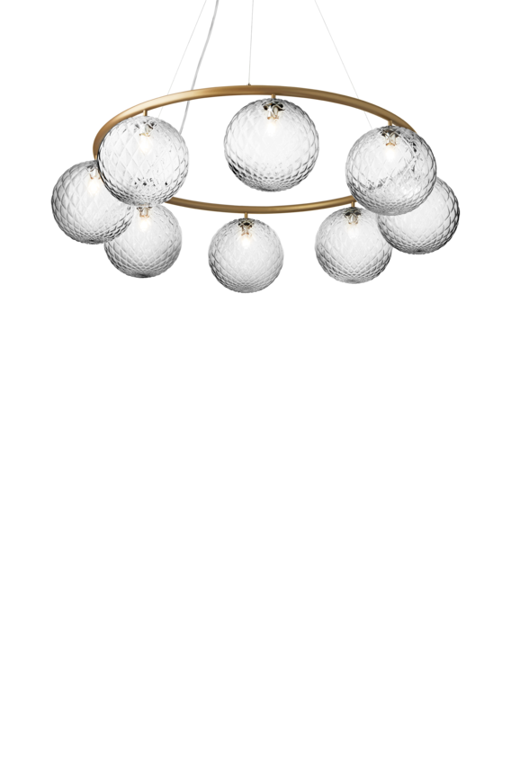 Lysekrone bestående af otte runde skærme i klart optikglas samt messingfarvet krone, på hvid baggrund