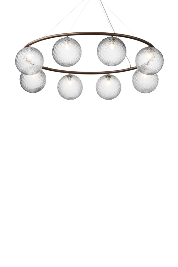 Lysekrone bestående af otte runde skærme i klart optikglas samt bruneret krone, på hvid baggrund