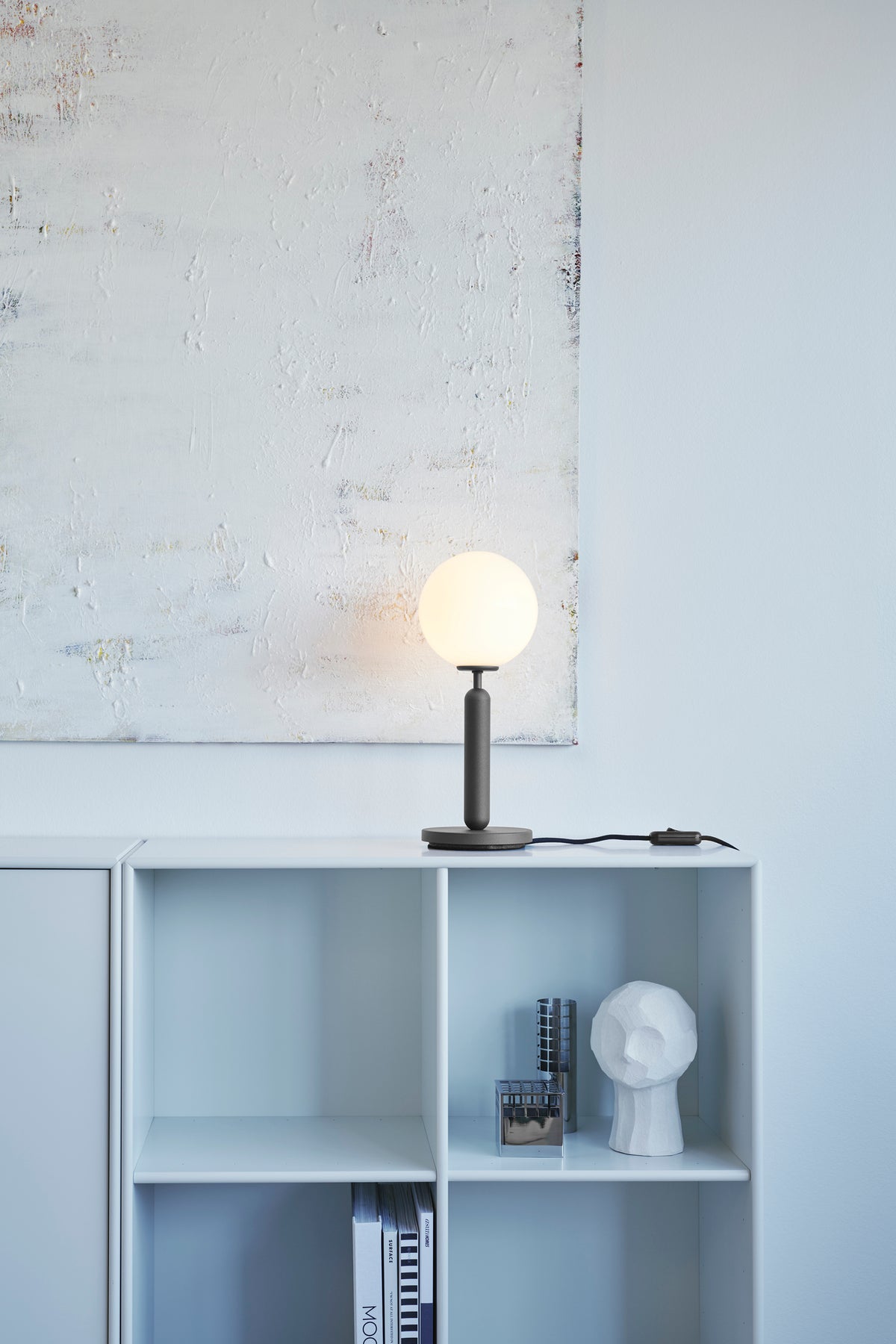 Bordlampe med rund skærm af opalglas og mørkegråt stel, på reol i dagligstuemiljø
