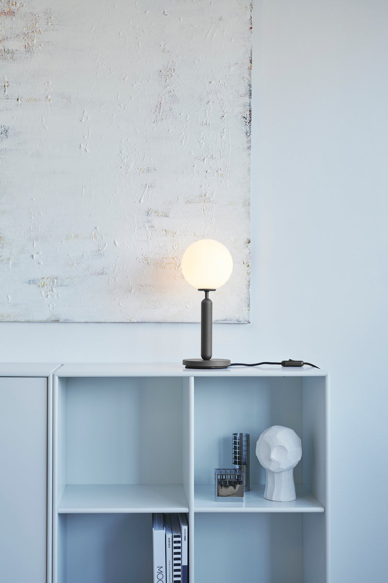 Bordlampe med rund skærm af opalglas og mørkegråt stel, på reol i dagligstuemiljø