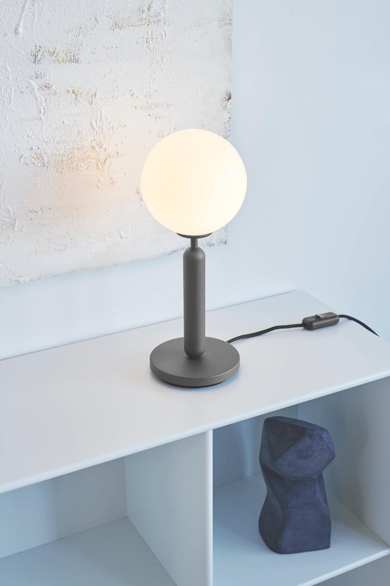 Bordlampe med rund skærm af opalglas og mørkegråt stel, på reol i dagligstuemiljø