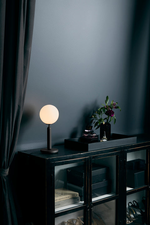 Bordlampe med rund skærm af opalglas og mørkegråt stel, på vitrine i dagligstuemiljø