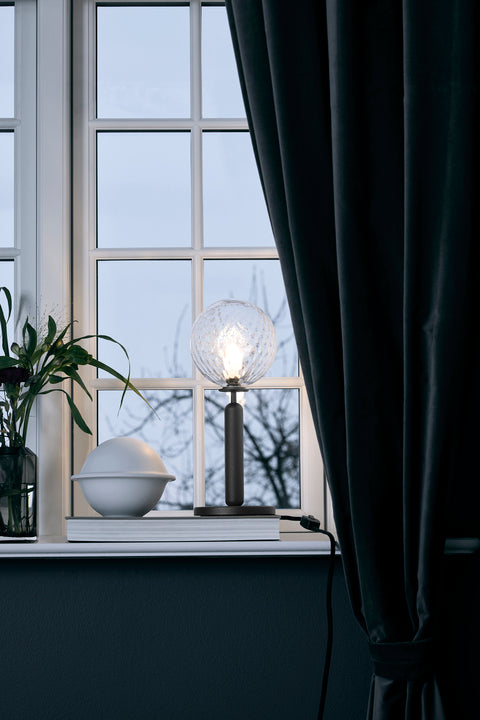 Bordlampe med rund skærm af klart optikglas og mørkegråt stel, i dagligstuemiljø i vindueskarm