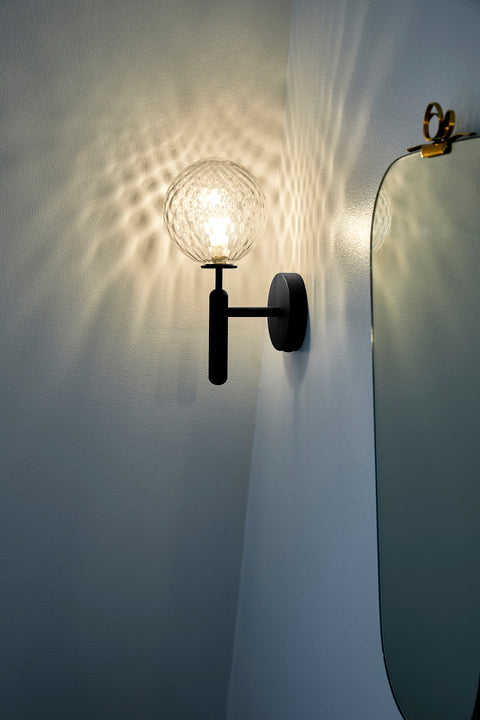 Væglampe med rund skærm af klart optikglas og mørkegråt stel, på hvid væg ved siden af spejl