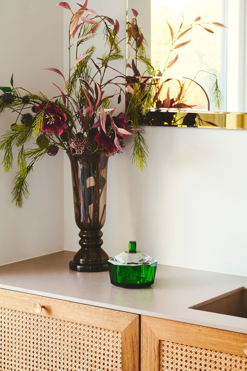 Badeværelsesmiljø med bonbonniere i klar og grøn krystal, stående ved siden af vase med blomster på badeværelsesmøbel i træ.