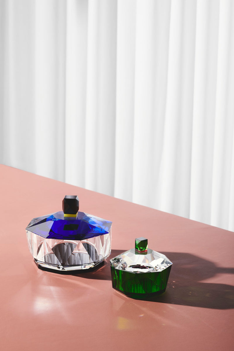 To bonbonnierer i klar, grøn og blå krystal, på rosafarvet bord og med hvidt gardin i baggrunden.