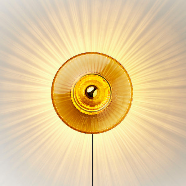 Rund væglampe i amber (orange/gul) plisseret glas, som skaber et lysspil på væggen. 