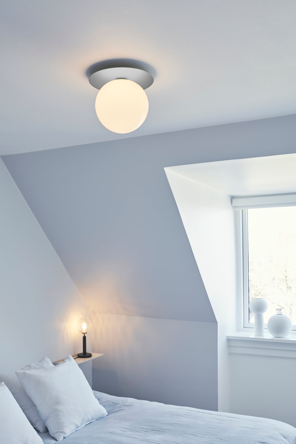 Bordlampe med rund skærm af klart optikglas og mørkegråt stel, ved siden af seng i soveværelse