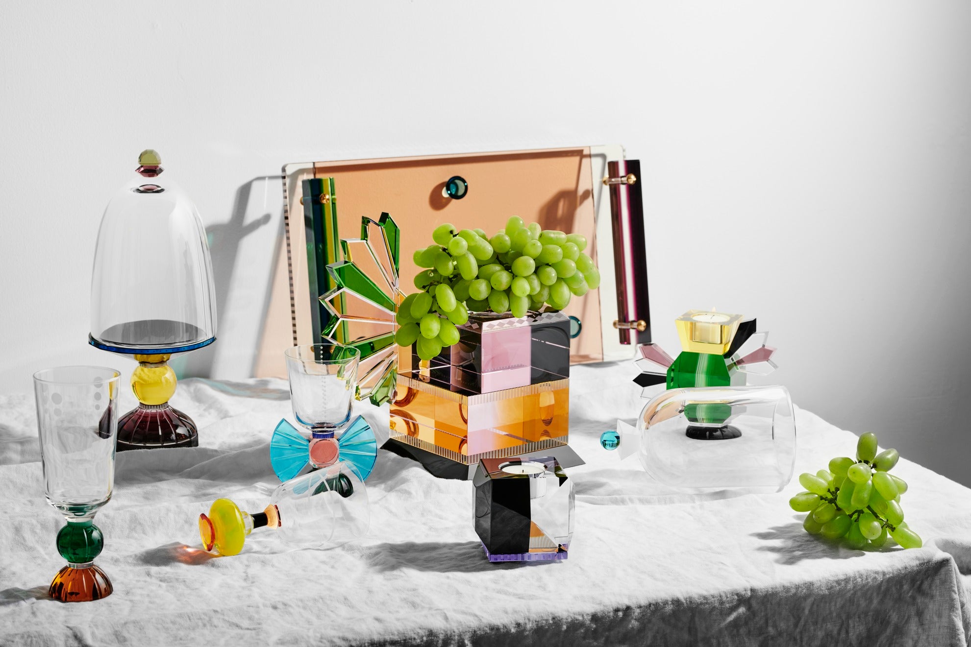 Opstilling af forskellige lysestager, glas  og skåle i farvet krystal, på middagsbord med dug og frugt.
