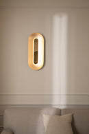Oval messingfarvet væglampe med indirekte lys, i dagligstuemiljø