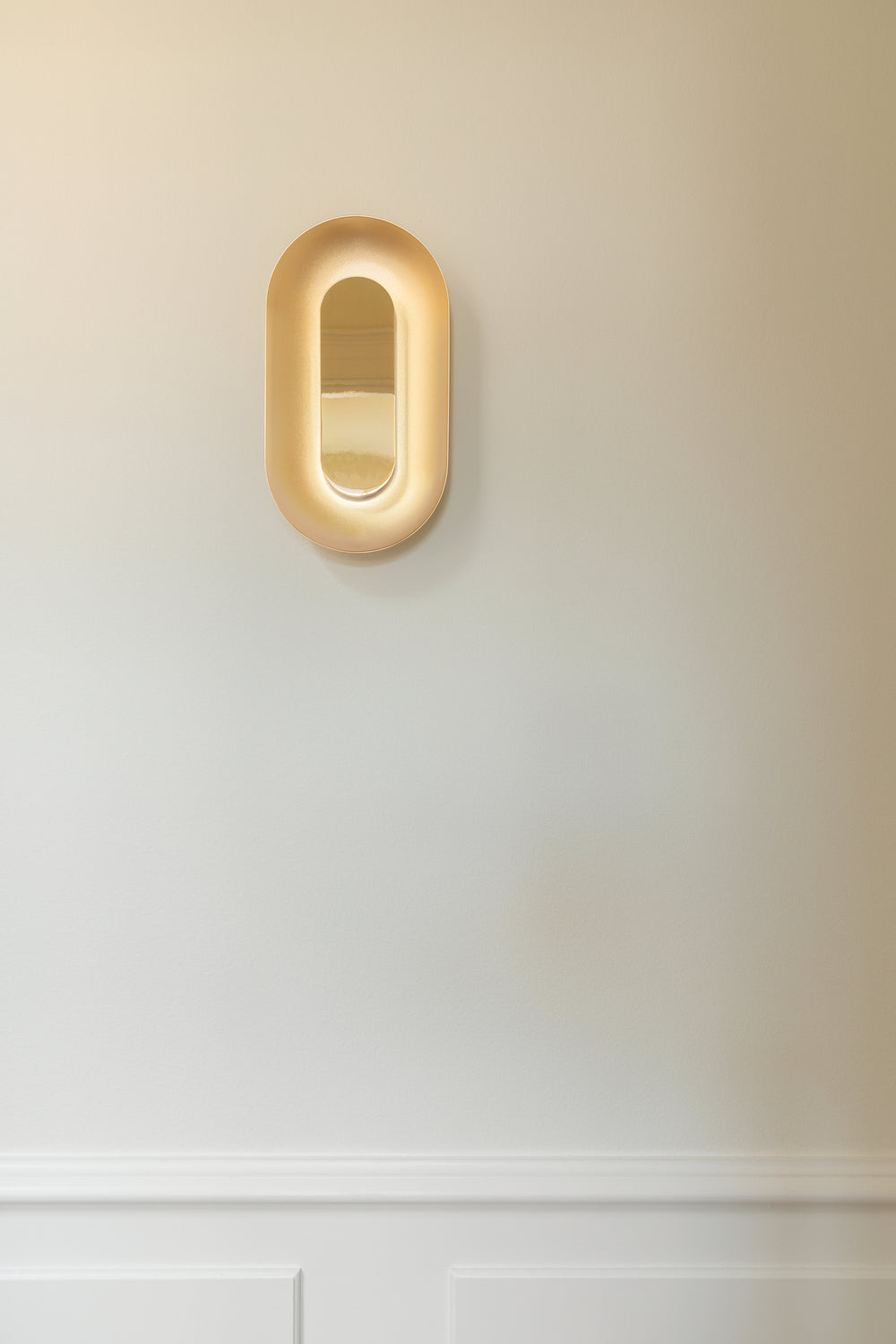 Oval messingfarvet væglampe med indirekte lys, på hvid væg