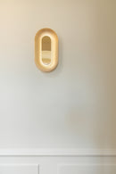 Oval messingfarvet væglampe med indirekte lys, på hvid væg