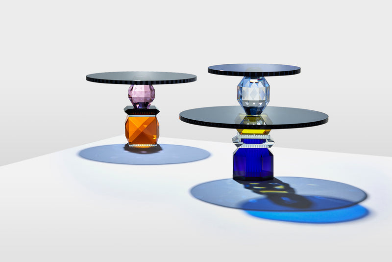 Opstilling af to forskellige opsatser i farvet krystal, på hvid bord med lysegrå baggrund.