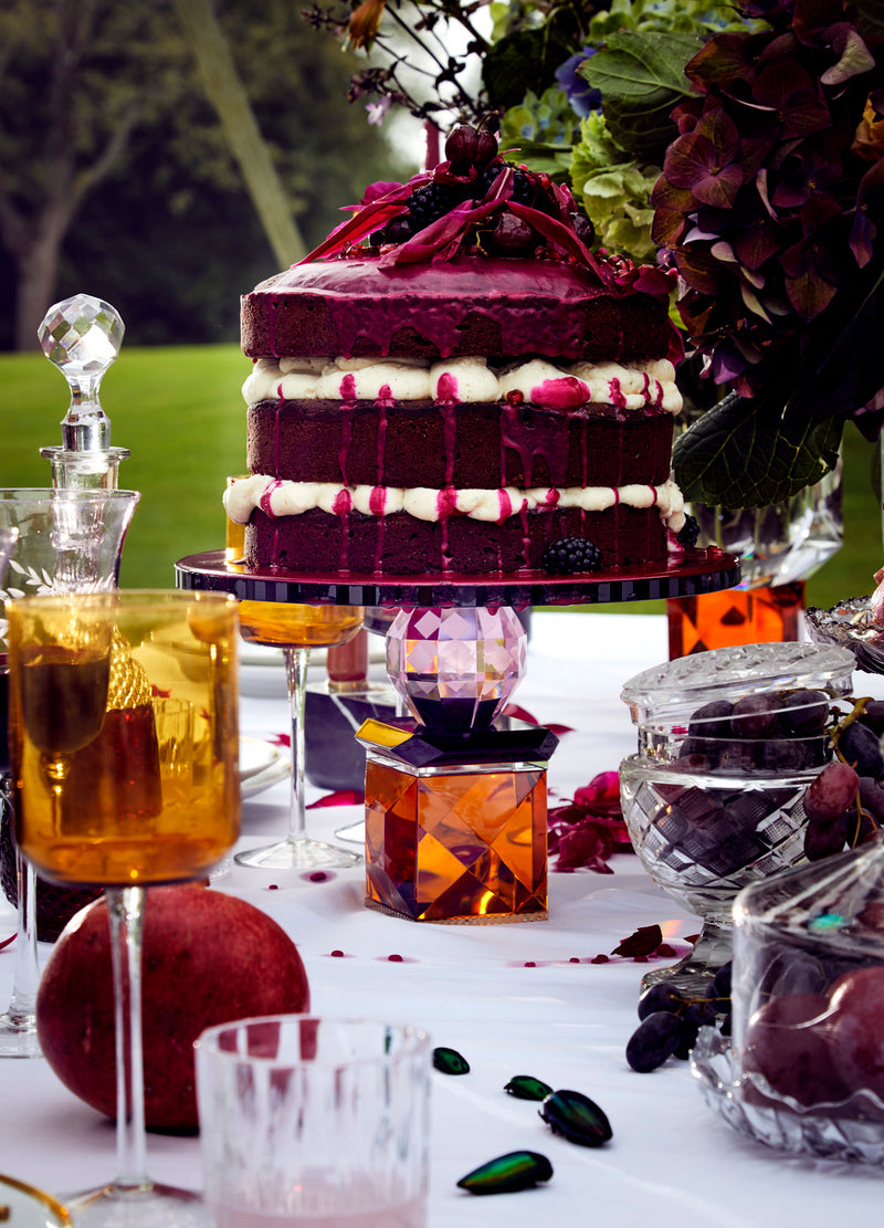 Lagkage på kagefad af farvet krystal stående på bord dækket med krystalglas samt fugter.