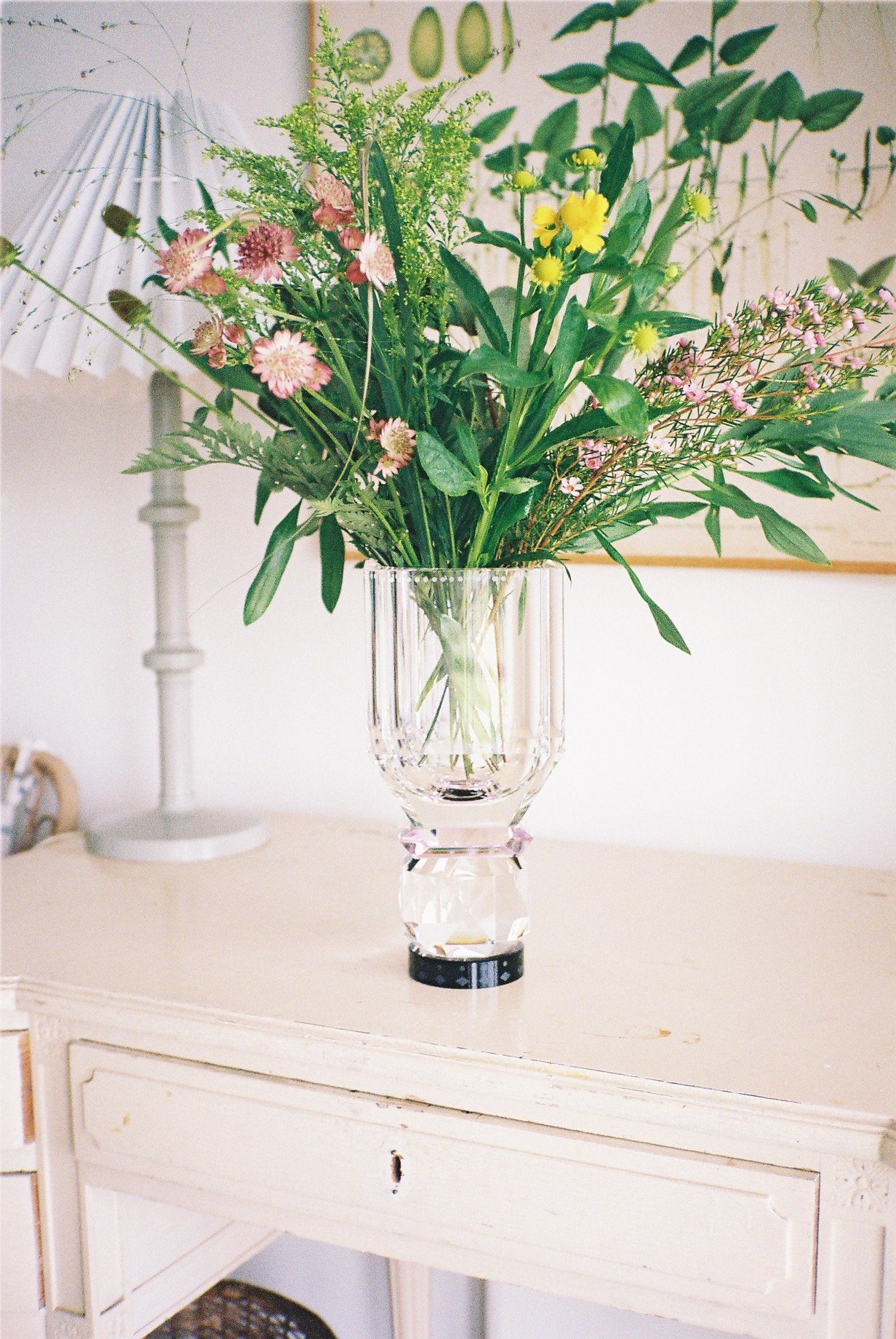 Vase i klar, lyserød og sort krystal med blomster i, stående på hvidt bord med lampe og kunst i baggrunden.