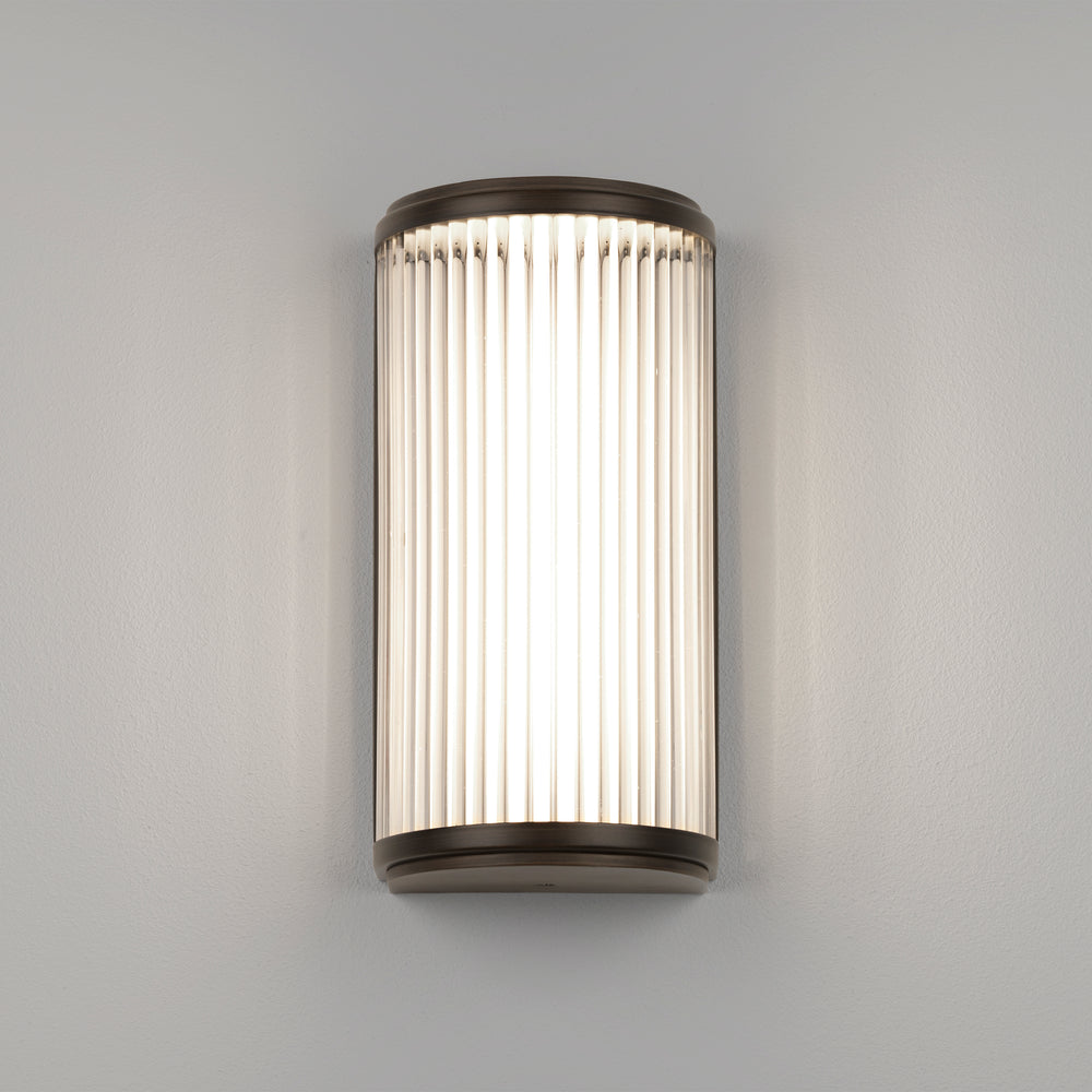 Lampe med top, bund og bagside i bronzefarvet stål samt en glasskærm med nedadgående linjer.