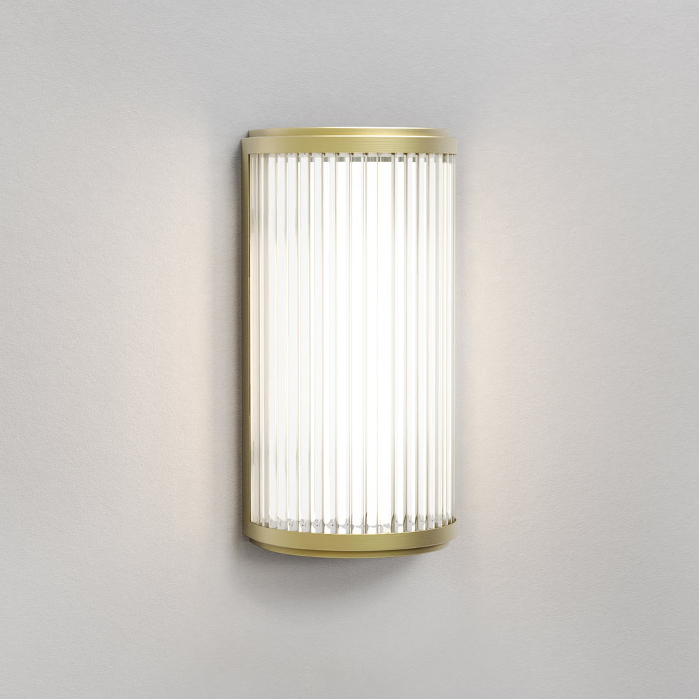 Lampe med top, bund og bagside i guldfarvet stål samt en glasskærm med nedadgående linjer.