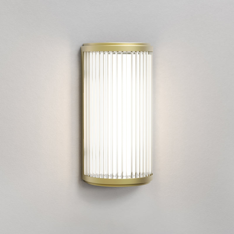 Lampe med top, bund og bagside i guldfarvet stål samt en glasskærm med nedadgående linjer.